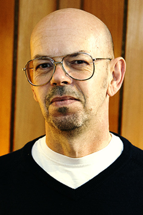 Porträtfoto von Ulf Treger, frontal, im blauen V-Neck vor einer Holzwand